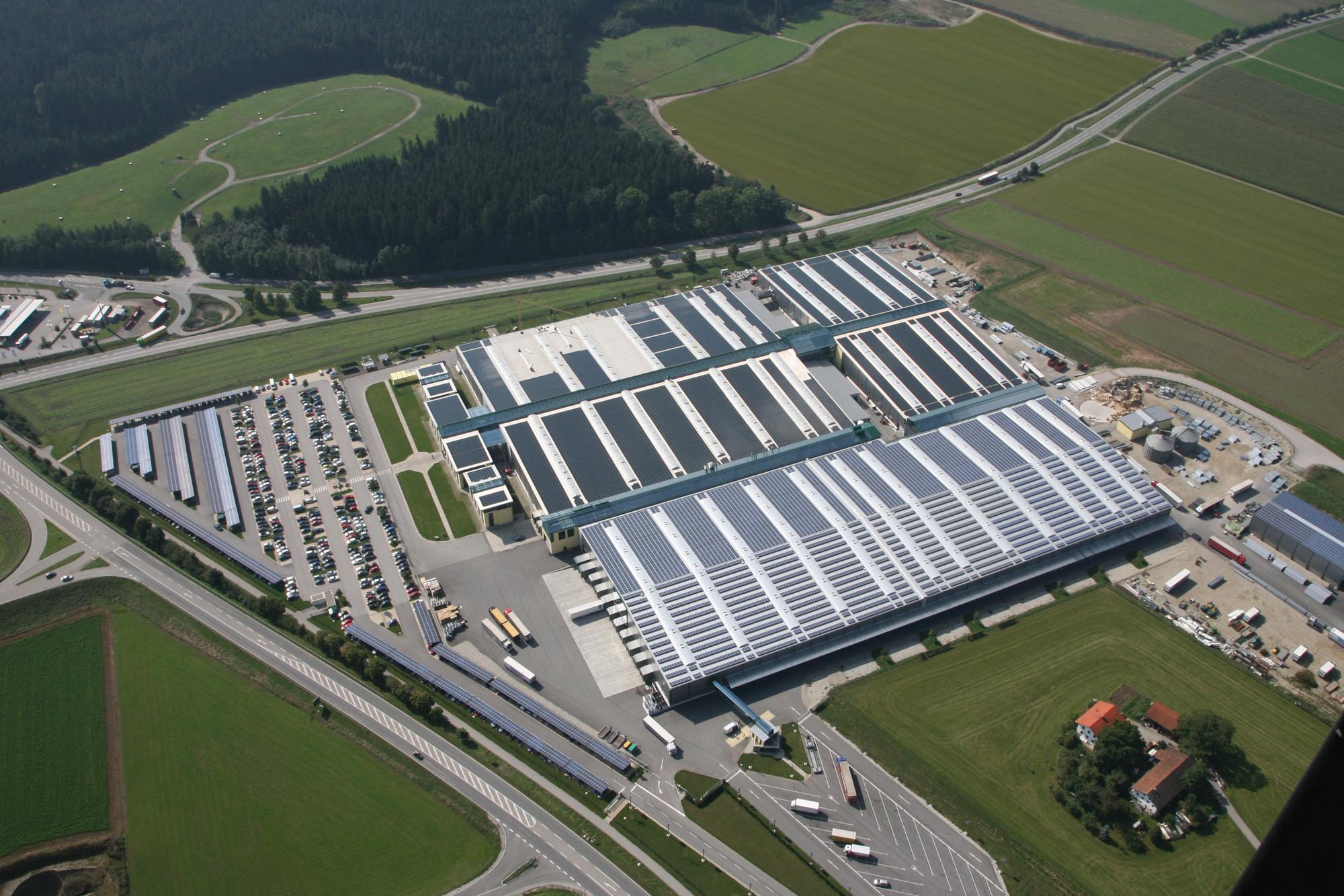Aerial view of a large industrial facility with adjacent parking lots and surrounding green fields. Multiple buildings with white and blue rooftops are visible. Nearby roads and a small house are also shown.